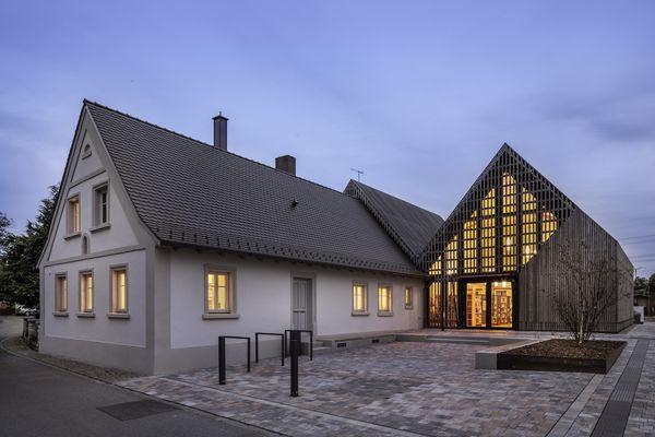 Die Bücherei in Gundelsheim im Abendlicht. Die Fenster sind hell erleuchtet. im Vordergrund ist das alte, sanierte Stallgebäude. Im Hintergrund ein Neubau aus Holz und Glas, der die Dachform aufnimmt.