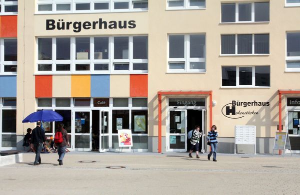 Bürgerhaus in Hohenstücken, Brandenburg (beiger Neubau, ausschnittsweise sichtbar). Zwei Kinder laufen aus dem Eingang und zwei Erwachsene gehen auf ihn zu. Weitere Personen sitzen unter einem blauen Sonnenschirm vorm Café.